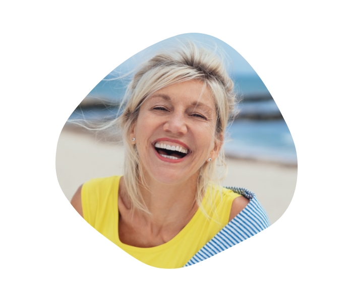 Bild einer Frau mittleren Alters am Strand, die fröhlich in die Kamera lacht.