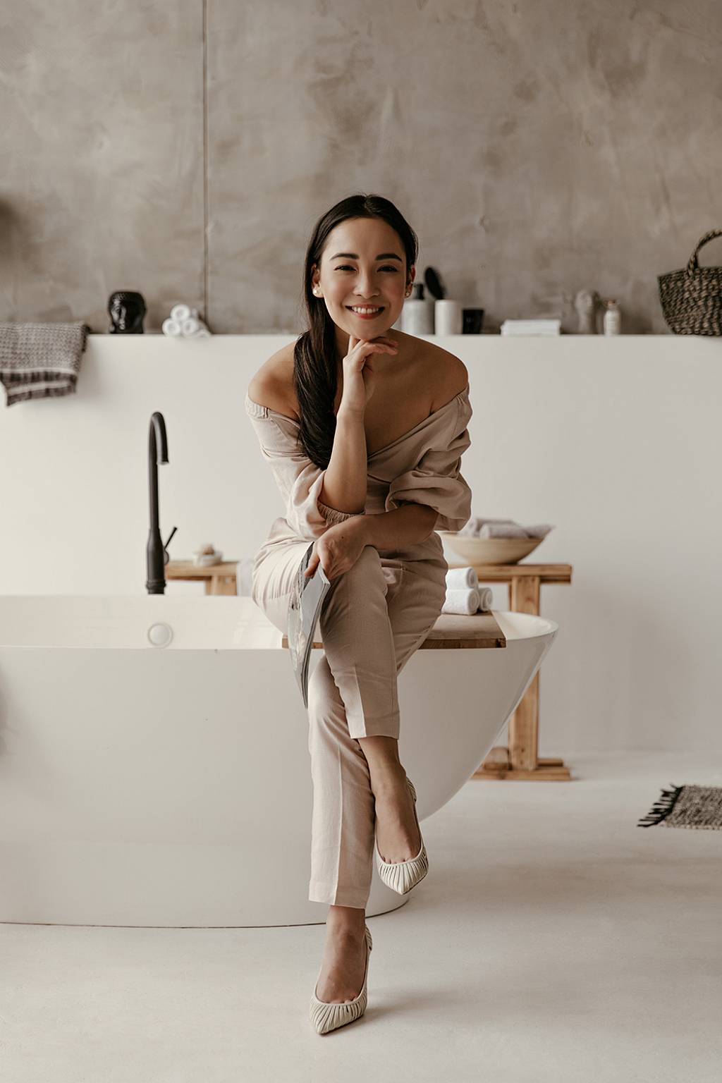 Junge Frau mit langen Haaren sitzt in einem beigen Kostüm auf dem Rand einer Badewanne und lächelt, das Kin auf Ihrer Hand aufgestützt, in die Kamera. Das Badezimmer selbst ist modern und schlicht eingerichtet.