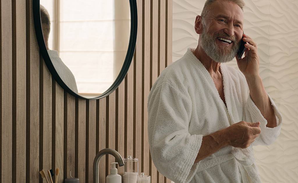 Älterer Mann in einem weißen Bademantel steht, angelehnt an seinen Waschtisch, in seinem Bad und telefoniert freudig mit seinem Smartphone.
