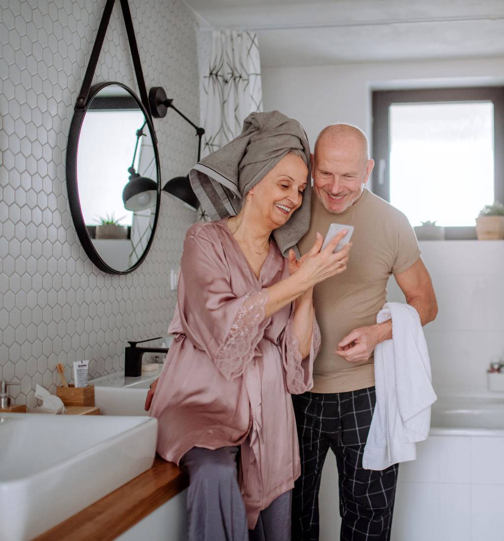 Älteres Paar steht lächelnd im Badezimmer und schaut gemeinsam auf ein Smartphone; die Frau trägt einen Bademantel und hat ein Handtuch um den Kopf gewickelt, der Mann hält ein Handtuch in der Hand.