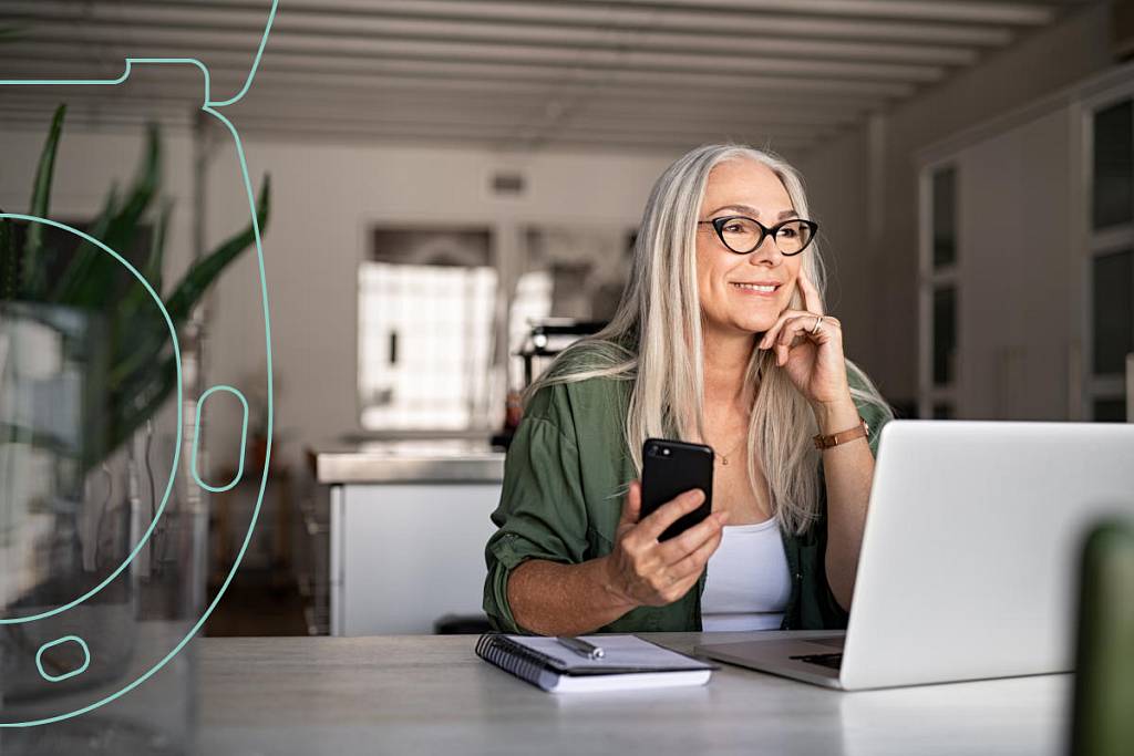 Ältere Frau mit langen grauen Haaren und Brille sitzt lächelnd an einem Tisch mit Laptop, Smartphone und Notizbuch; links im Bild ein transparenter Umriss des EKG-seats mit Sensorflächen.