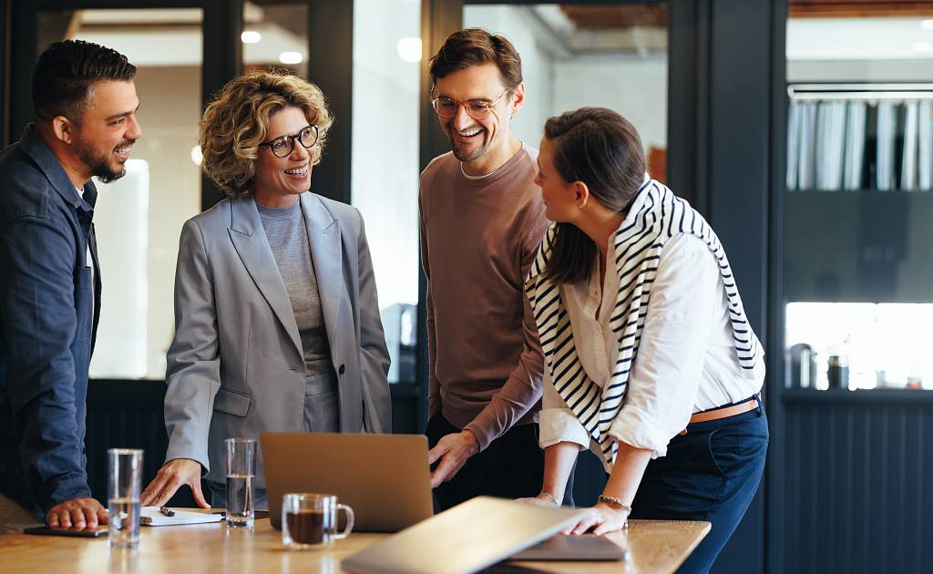 Vier Personen stehen lachend um einen Tisch in einem modernen Büro und schauen auf einen Laptop – sie wirken engagiert und im Gespräch vertieft.
