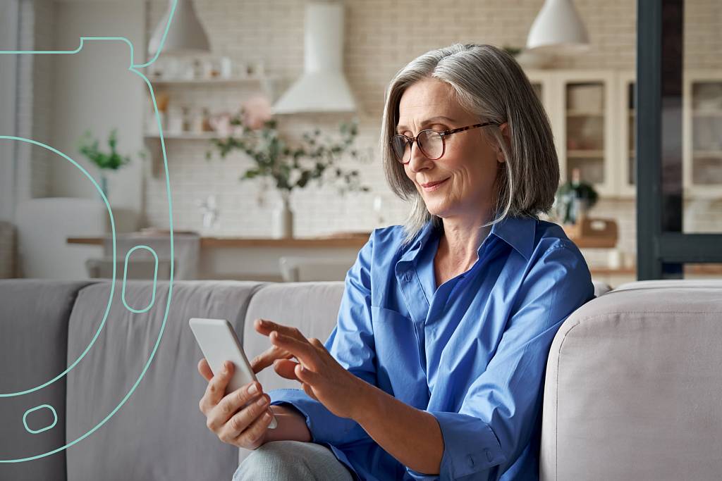 Ältere Frau mit grauen Haaren und Brille sitzt auf einem Sofa und benutzt ein Smartphone.